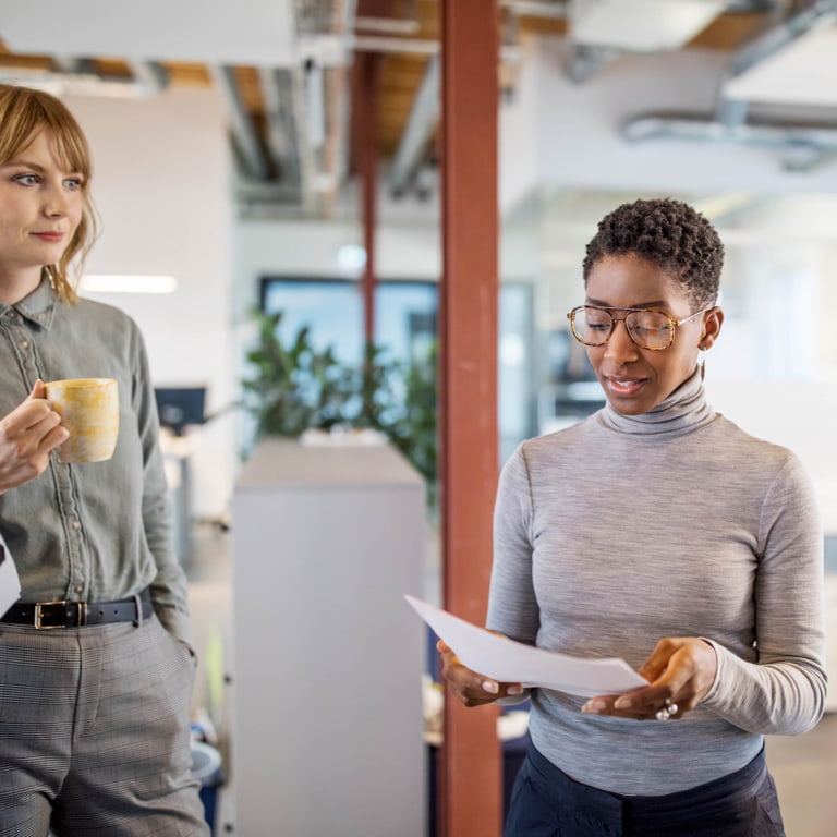 Two women standing in a modern open plan office having a discussion about a few documents.