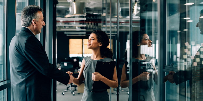 A business man and women in black corporate clothing shaking hands at a hallway of a modern glass office building.