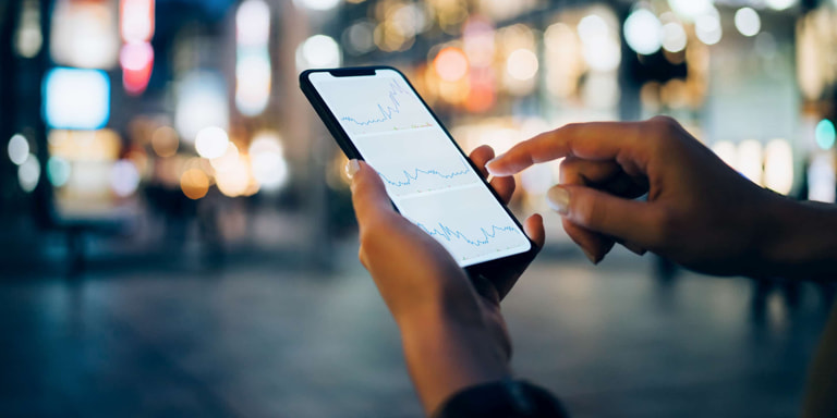 A man on a busy city street at night, looking at share price movements on his cellphone.