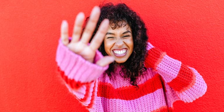 A young woman in a vibrant striped pink and red jersey, standing against a red background, holding out her hand and partially closing her eyes.