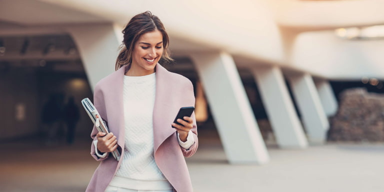 A woman standing behind a building smiles while looking at her cell phone and holds a file in her other hand.