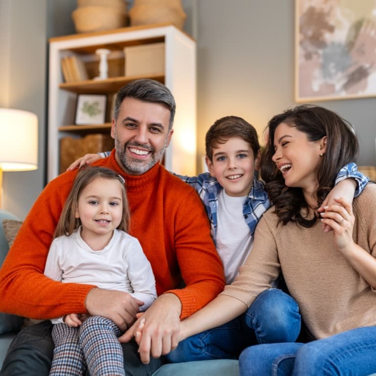 Four people sit on a couch in a cosy living room, smiling at the camera. The background has lamps, framed pictures, and shelves with decorative items.