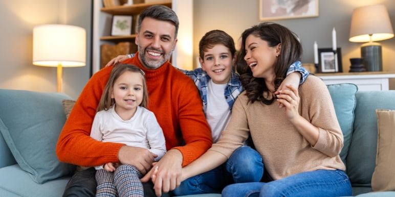 Four people sit on a couch in a cosy living room, smiling at the camera. The background has lamps, framed pictures, and shelves with decorative items.