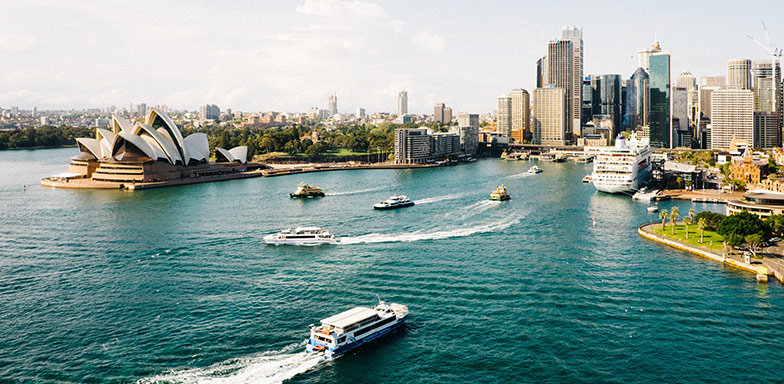 A view of the Sydney Opera House surrounded by a deep blue sea and the Sydney city skyline. 
