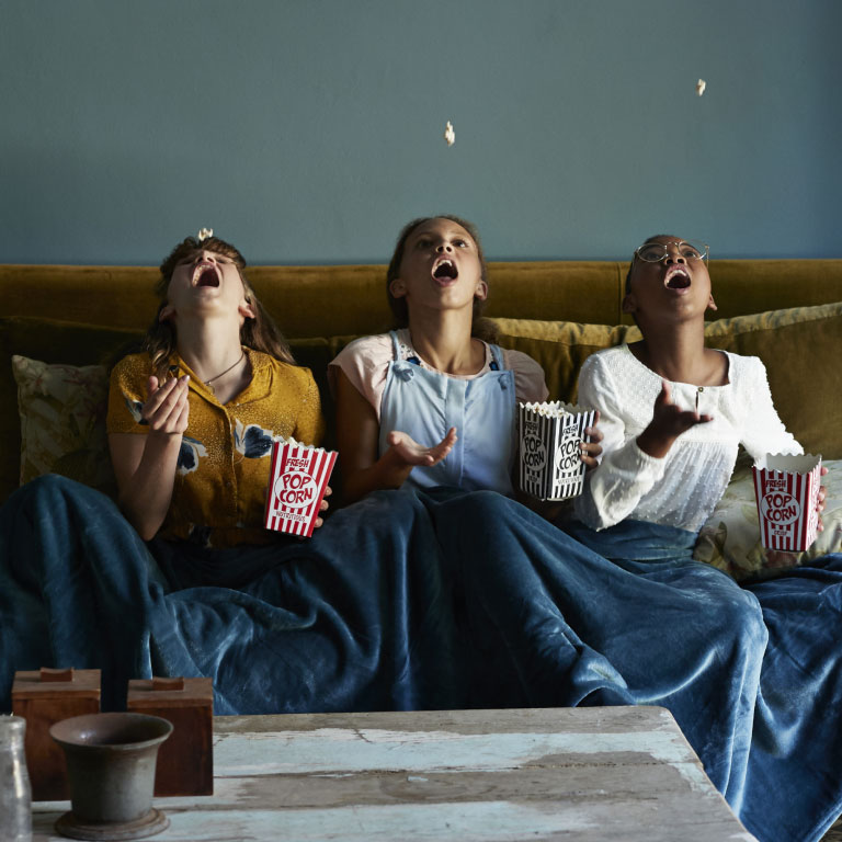Three girls sitting and eating popcorns on a couch with a blanket in a home bought with the help of FundsAtWork's housing loan guarantee.