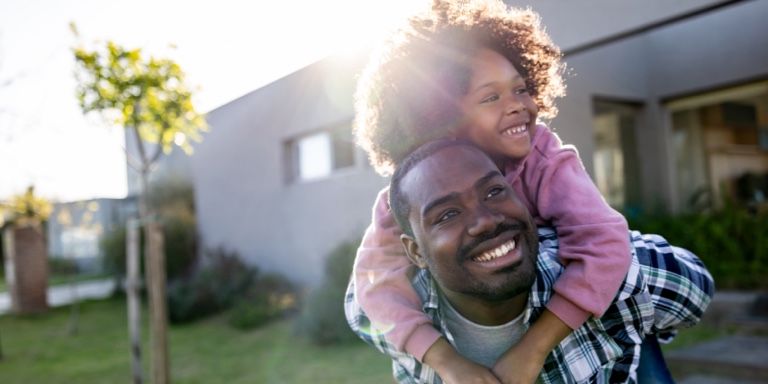 A dad holding his daughter on his back and shoulders in their backyard.