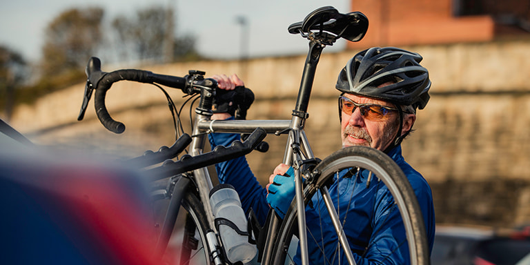 An older man wearing his cycling head gear busy mounting his bicycle on the car.