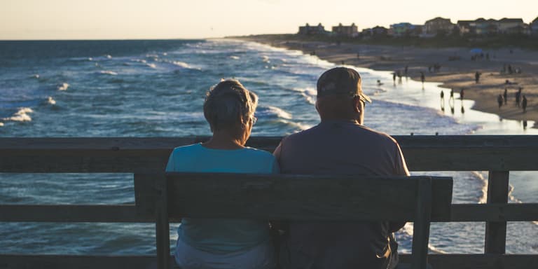 A view of an older couple from behind, sitting on a bench looking out at the waves gently crashing on the shore and people walking on the beach.