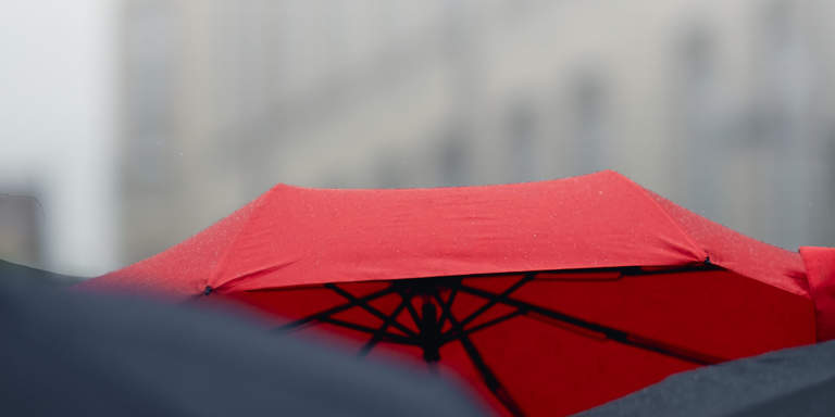 Red umbrella over many black umbrellas against a grey, cloudy sky.