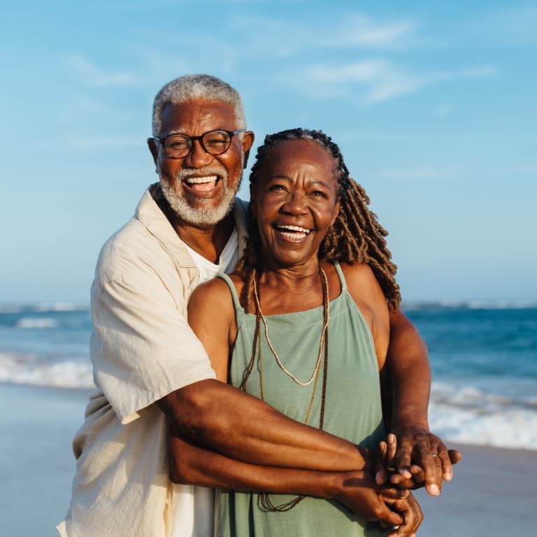 Two people stand on a beach, smiling and embracing. The ocean stretches behind them under a clear blue sky, creating a peaceful and joyful scene.