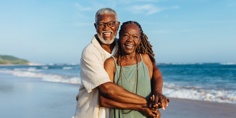 Two people stand on a beach, smiling and embracing. The ocean stretches behind them under a clear blue sky, creating a peaceful and joyful scene.