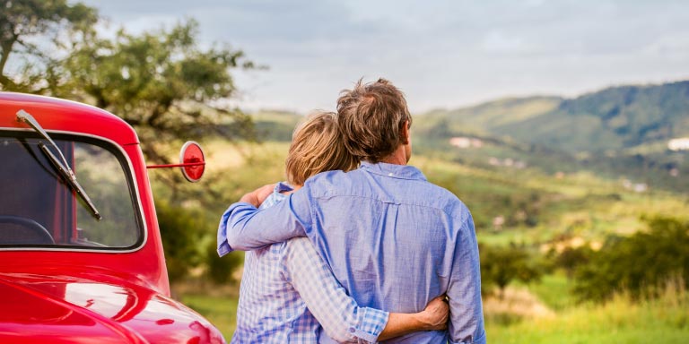 Senior couple hugging next to a red vintage car looking at green rolling hills.