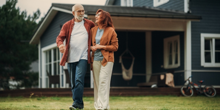 Two people walk arm-in-arm on a lawn in front of a blue house with white trim. A bicycle and hammock are visible in the background, suggesting a relaxed, homey setting.