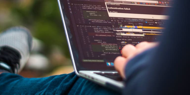 A man in jeans and a sweater, sitting on the grass outside, working on a laptop.