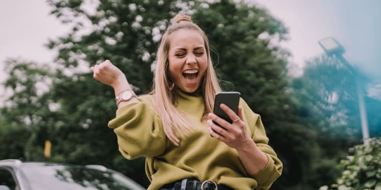 A young woman leaning against her car looking at her cell phone excitedly as she seamlessly activated her Safety Returns.