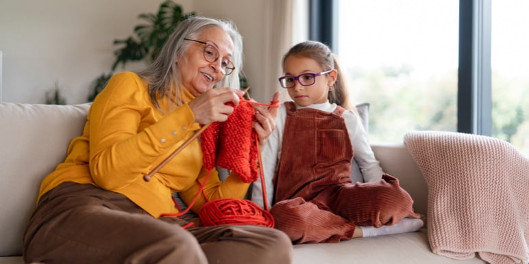 A retired woman sitting on the couch with her granddaughter teaching her how to knit. 