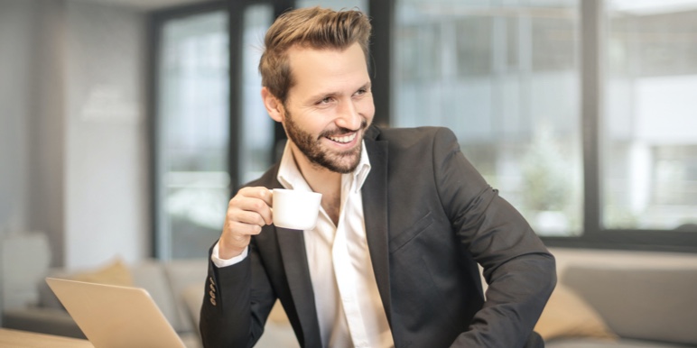 A confident young man seated at a desk with his laptop drinking his cappuccino.