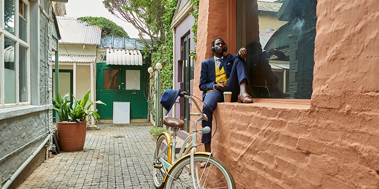 A man in a suit sitting by the window and waering headsets with his bicycle parked next to him.