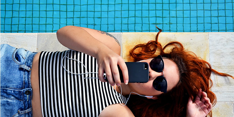 A red-haired lady in shades lying next to a pool and taking a selfie.