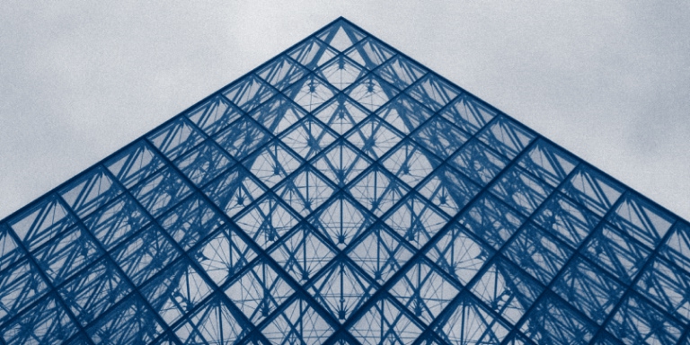 Top portion of a glass and steel pyramid structure - the design features a geometric grid of triangular glass panels framed by metal beams, creating a symmetrical pattern that converges to a point at the top.