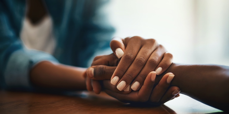 A woman in a denim shirt and white vest wrapping her hands around someone else's hand to comfort them.