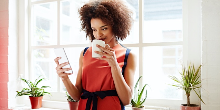 A woman in a red shirt, with a coffee mug in her hand, standing next to a glass wall while typing  on her cell phone.