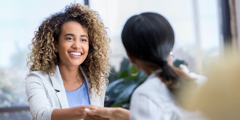 Two women smiling at each other and shaking hands inside an office building.