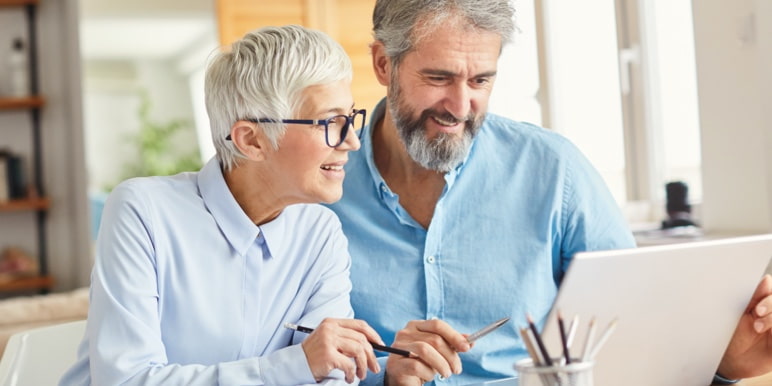 A middle aged couple with greying hair wearing shirts in different shades of blue holding pens while looking at their laptop screen.