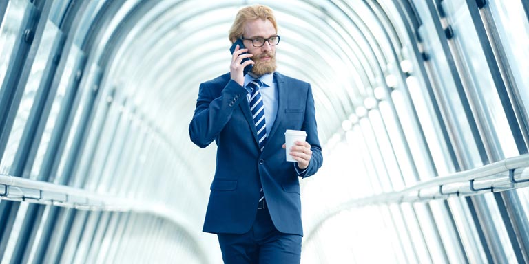 A min in a suit and tie, walking inside a modern office building, while talking on his cell phone.
