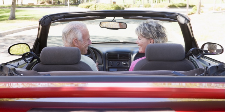 A back view of an older white couple with greying hair looking at each other smiling while driving down a road on a sunny day in their red convertible car.