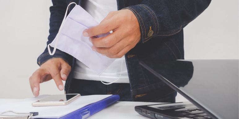 A young man wearing jeans, a dark denim jacket and a white t-shirt holding a mask in his left hand while he is typing on his cellphone that is on a desk alongside a half-open laptop and notebook.