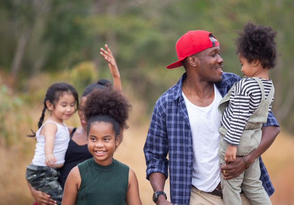 A family of five walking in a field with dad holding the little boy and mom holding the little girl while the older girl walks ahead of them. 