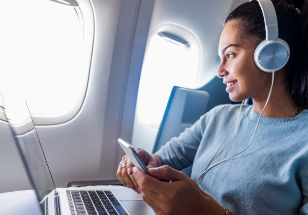 A lady sitting in a plan and looking through the window as she's listening to music on her headphones and working from her laptop.