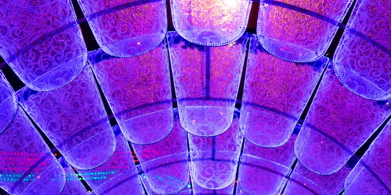 A colorful, decorative ceiling installation made up of rows of translucent, cylindrical panels glowing with blue and purple lights. The panels are arranged in an arched pattern and have intricate floral designs, creating a vibrant, immersive visual effect.