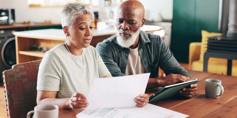 Two people sit at a table in a cosy kitchen. One holds a document, the other uses a tablet. They appear to be engaged in a focused discussion.