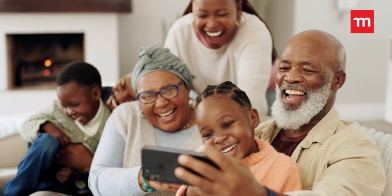 A multigenerational African family in a cozy living room, enjoying family time together and making memories by taking phone selfies. 