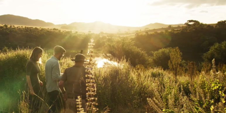A white man painting outside being watched by a white woman and a man.