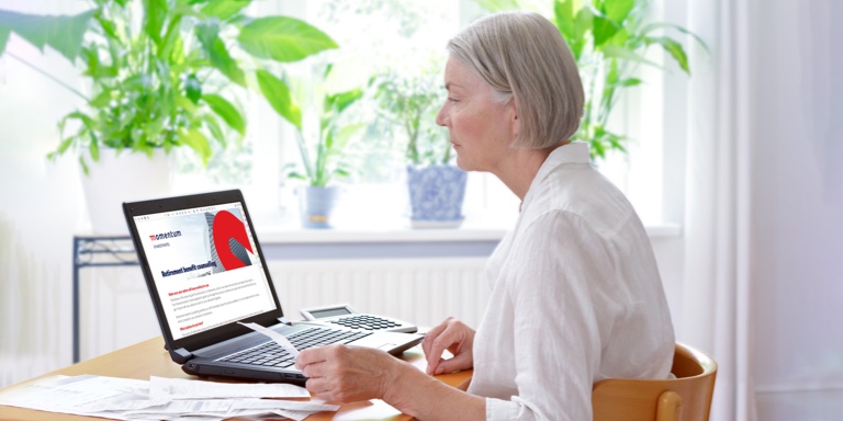 A white woman nearing retirement, seated at her home desk in front of a window with plants, applying for retirement benefit counselling on her laptop.