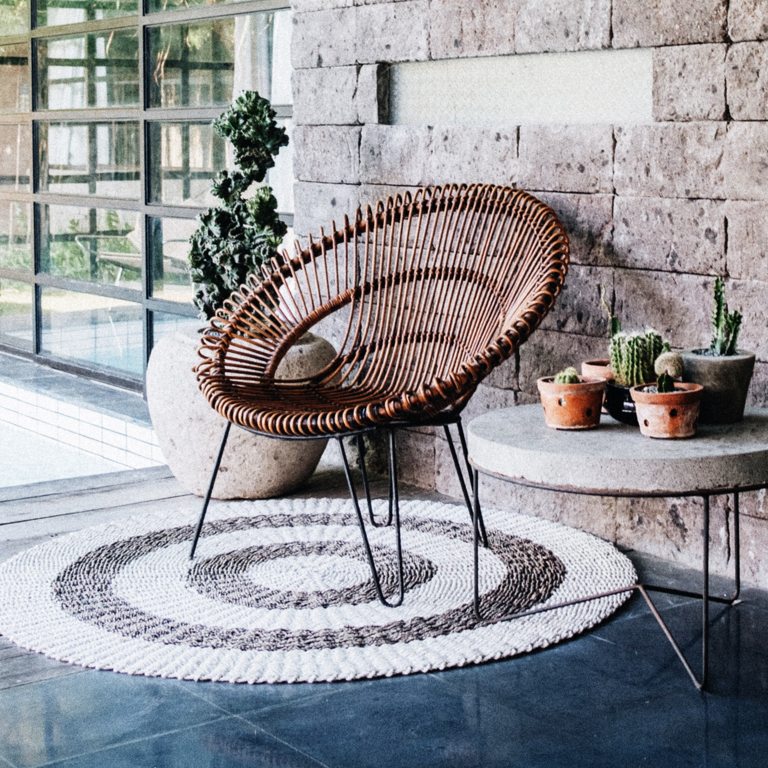 Cane chair on a grey and white coloured round carpet, next to a concrete table with metal legs that has a range of cacti plants displayed on it.