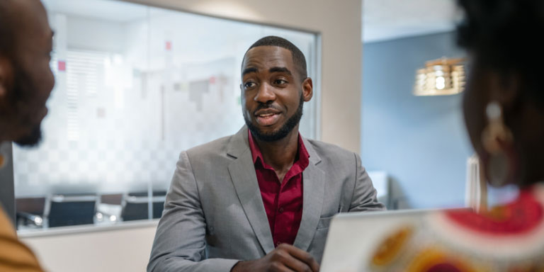 Man in a grey suit with maroon shirt talking to his colleagues
