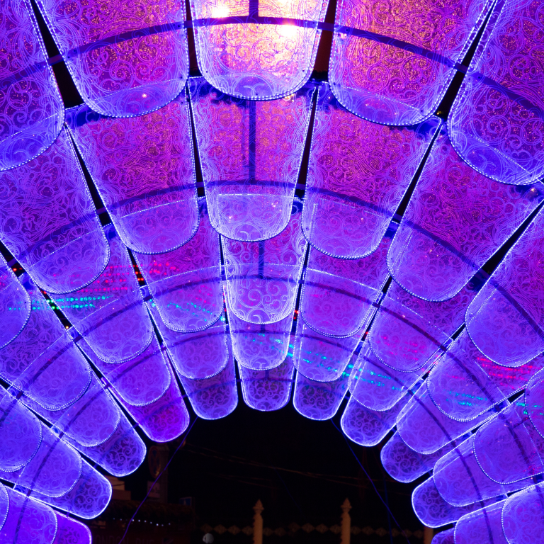 A colorful, decorative ceiling installation made up of rows of translucent, cylindrical panels glowing with blue and purple lights. The panels are arranged in an arched pattern and have intricate floral designs, creating a vibrant, immersive visual effect.